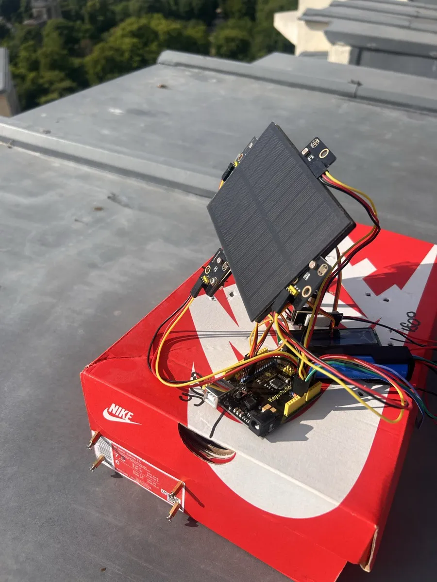 The solar tracker prototype on a rooftop: a small solar panel with light sensors on each side, mounted above an Arduino board and its wiring, on a red shoebox.